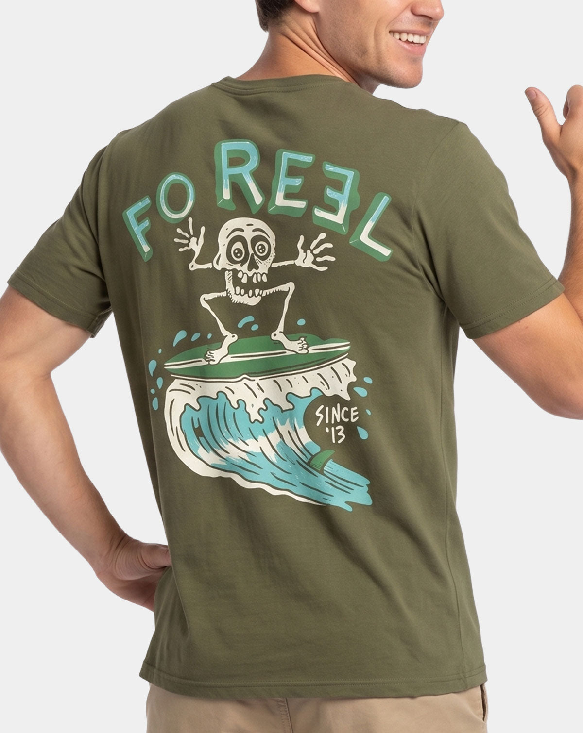 A man wearing an olive green t-shirt, shown from the back. The shirt features a large graphic print of a cartoon skull surfing on a wave with the text 'FO REEL' and 'SINCE '13'.