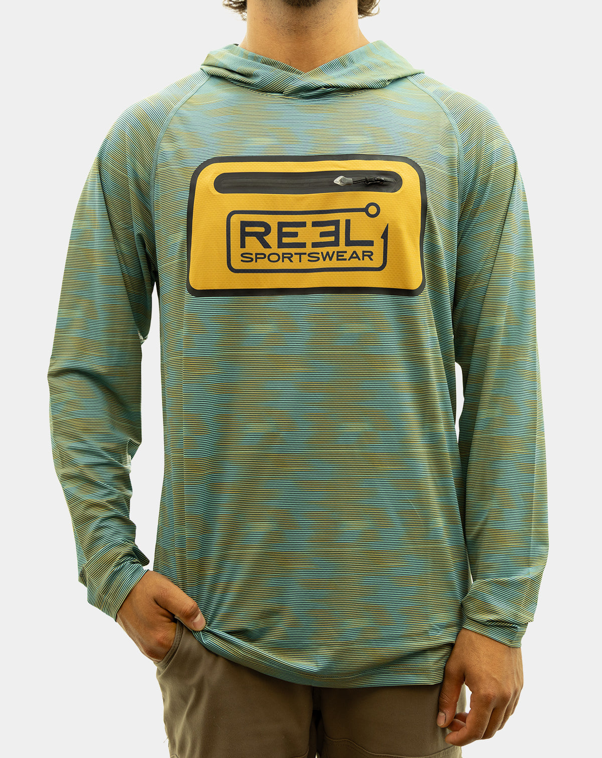 A man wearing the Static Pro+, a teal and olive green patterned long-sleeve hooded shirt with a yellow chest pocket featuring a black and blue 'REEL SPORTSWEAR' logo, paired with brown pants against a light gray background. fishing Shirt
