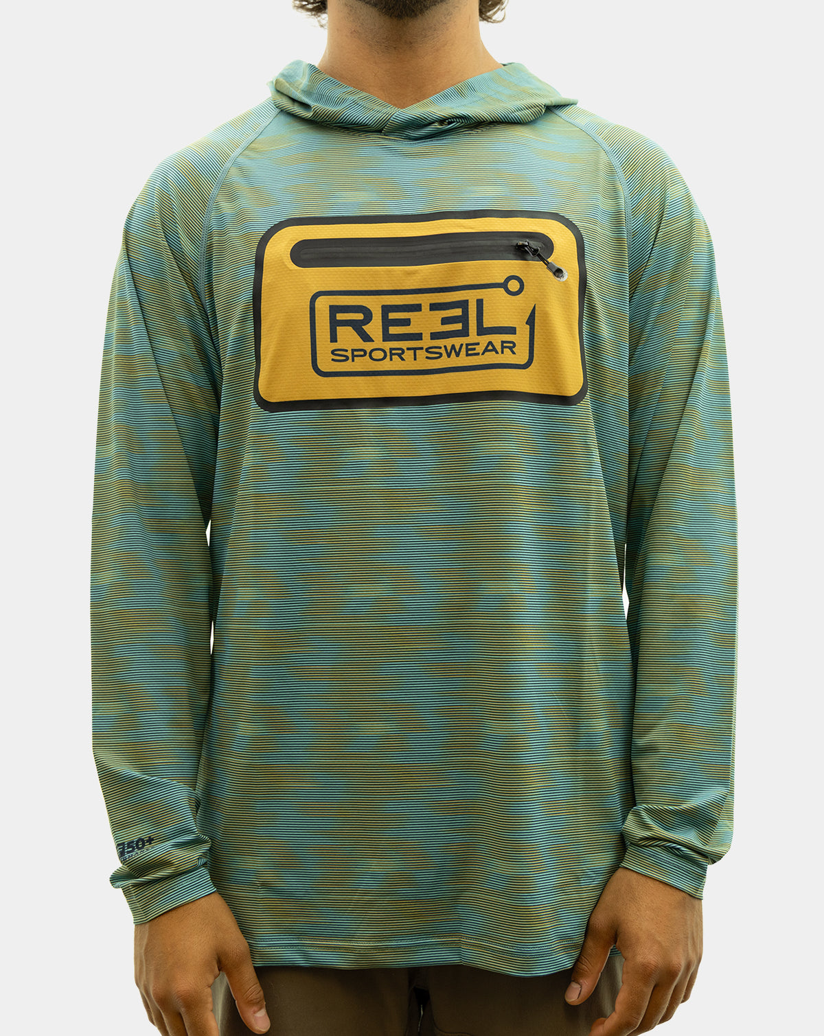A man wearing the Static Pro+, a teal and olive green patterned long-sleeve hooded shirt with a yellow chest pocket featuring a black and blue 'REEL SPORTSWEAR' logo, paired with brown pants against a light gray background. fishing Shirt