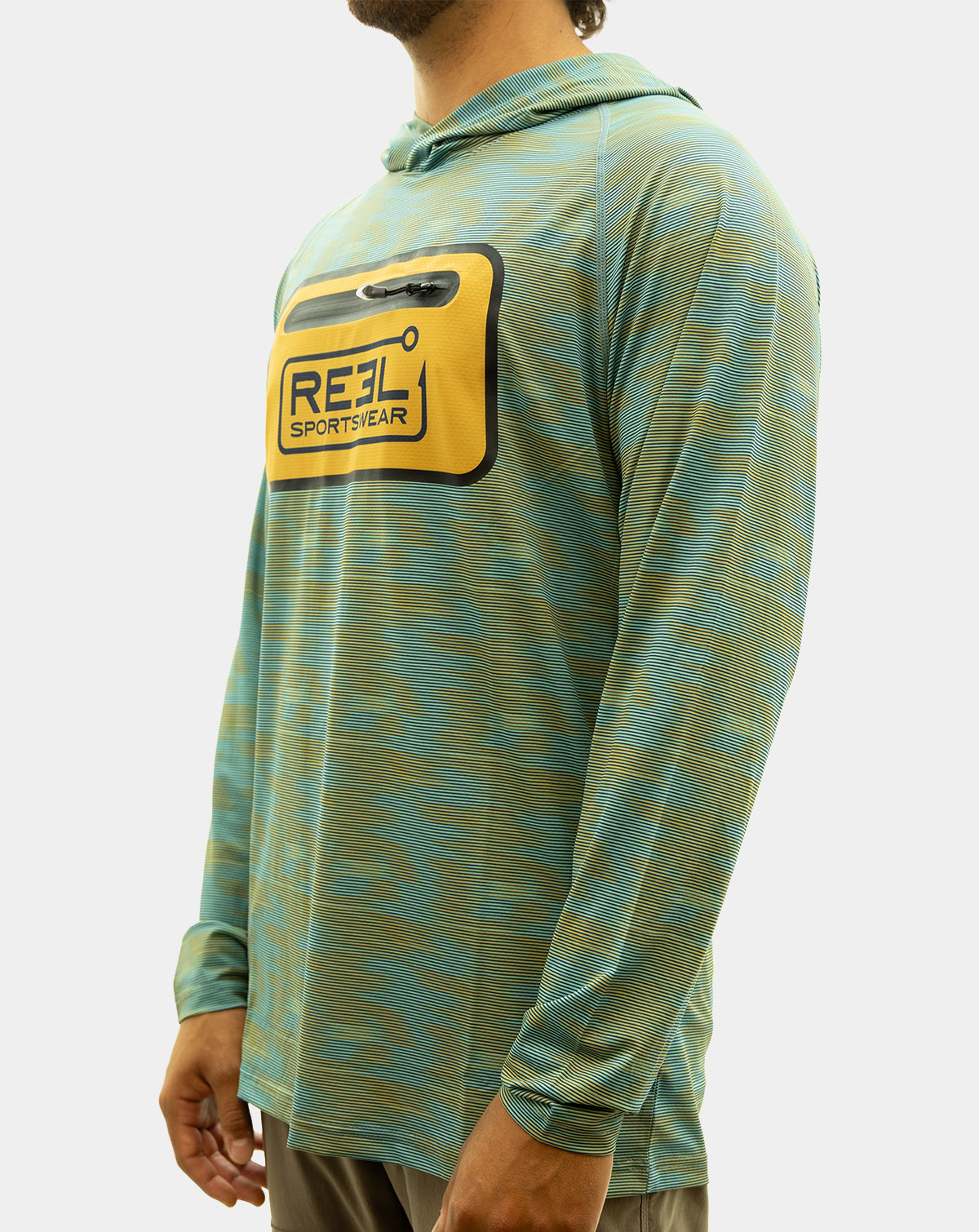 A man wearing the Static Pro+, a teal and olive green patterned long-sleeve hooded shirt with a yellow chest pocket featuring a black and blue 'REEL SPORTSWEAR' logo, paired with brown pants against a light gray background. fishing Shirt
