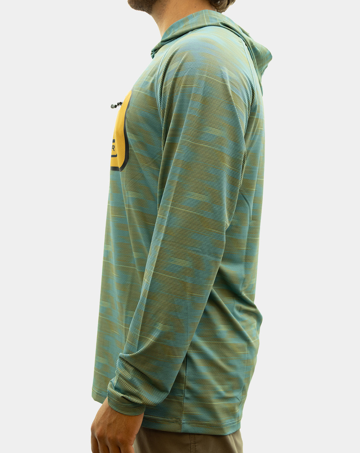 A man wearing the Static Pro+, a teal and olive green patterned long-sleeve hooded shirt with a yellow chest pocket featuring a black and blue 'REEL SPORTSWEAR' logo, paired with brown pants against a light gray background. fishing Shirt